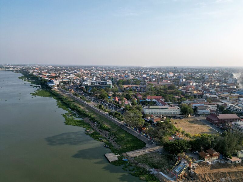 An aerial view of a low city of rooftops and roads stretching along the bank of a wide river under a pale sky.
