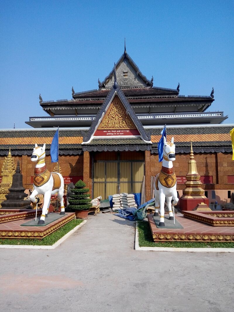 Two white horse statues on plinths in front of a Khmer temple hall with tiered roofs.
