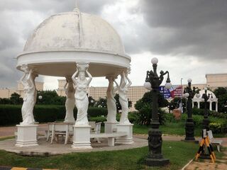A white domed pavilion held up by statues of women, in a park under a cloudy sky.