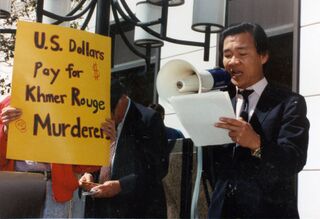 A man in a suit reads into a megaphone beside a sign that reads U.S. dollars pay for Khmer Rouge murderers.