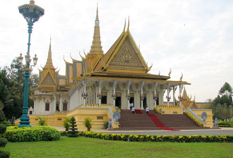 A cream and gold palace hall with tiered orange roofs and a tall gilded spire behind a red-carpeted staircase, with a green lawn and yellow flower beds in front.