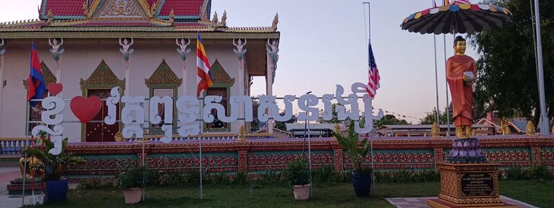 Ornate Khmer temple hall with red roof and garuda figures, white Khmer lettering sign in front and a standing Buddha statue under a parasol.
