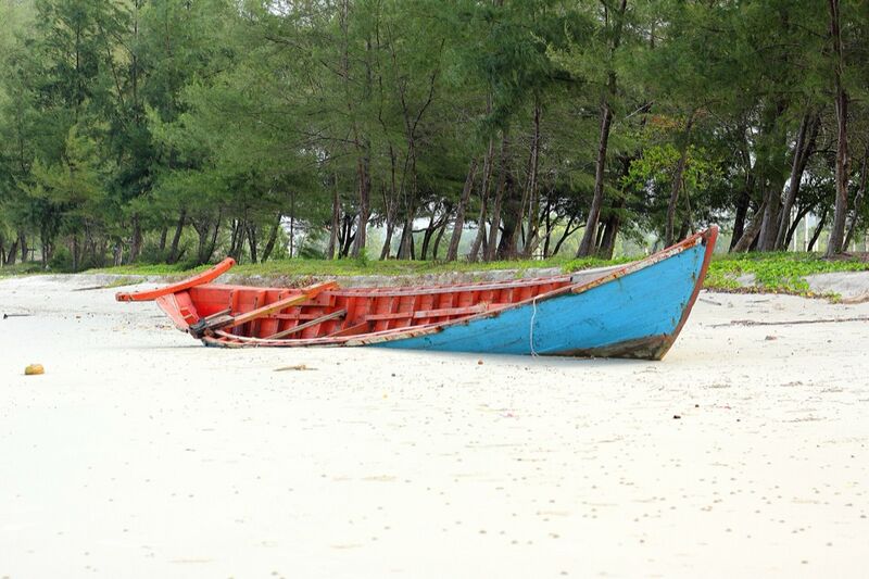 A red and blue wooden boat rests on white sand at the edge of a pine-lined beach, with no people in view.