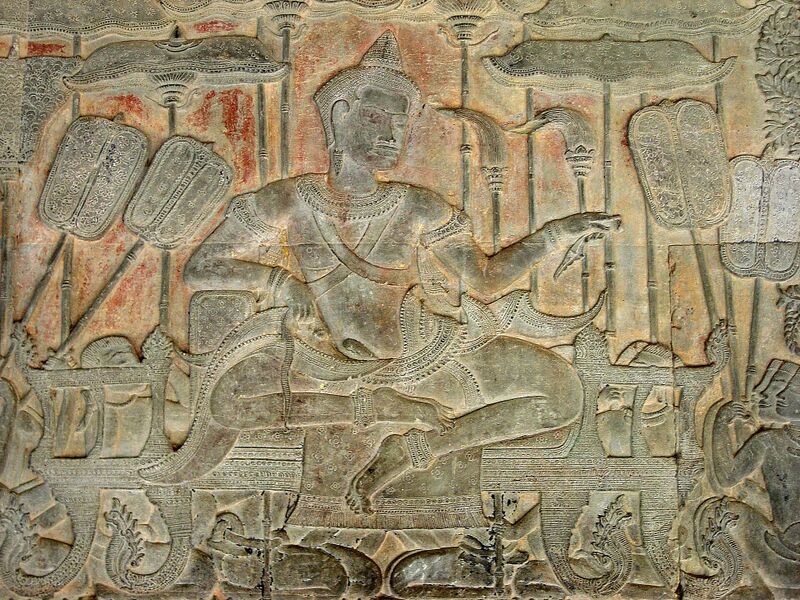 A sandstone relief of a crowned man seated cross-legged on a low throne, one arm stretched forward, under carved parasols and beside tall oval fans, with traces of red paint on the wall behind him.