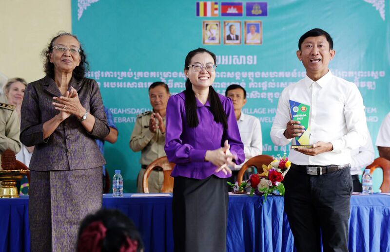 Three people standing at a ceremony stage, one holding an award, in front of a blue banner and seated guests.
