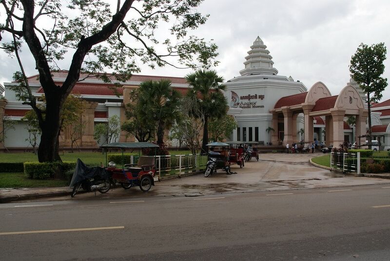 A long cream building with red tiled roofs and a tiered white tower bearing the museum's name in Khmer and English. Pink stone arches frame the entrance, with palm trees, tuk-tuks and a wet road in front under a gray sky.