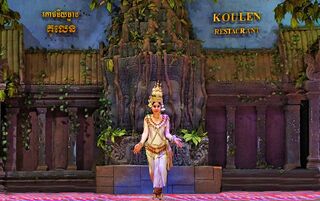 A solo dancer in a crown and embroidered costume performs on a stage set designed like a temple wall.