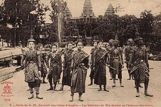 A line of dancers in crowns and sbai performing in front of the towers of Angkor Wat.