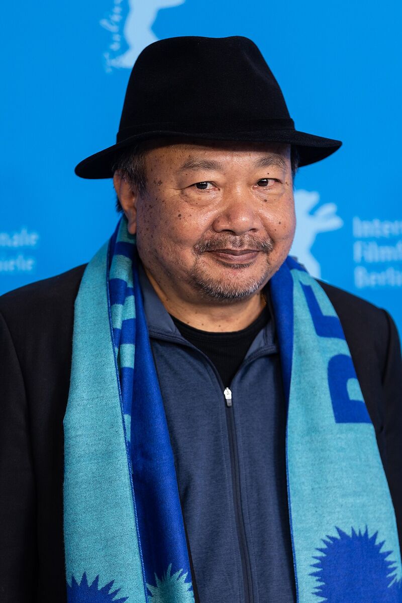 A man in a black hat, dark jacket and blue patterned scarf smiling in front of a blue festival backdrop.