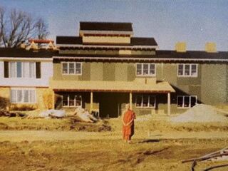 Older colour photo of a two-storey house under renovation, a monk in an orange robe standing on the bare earth in front.
