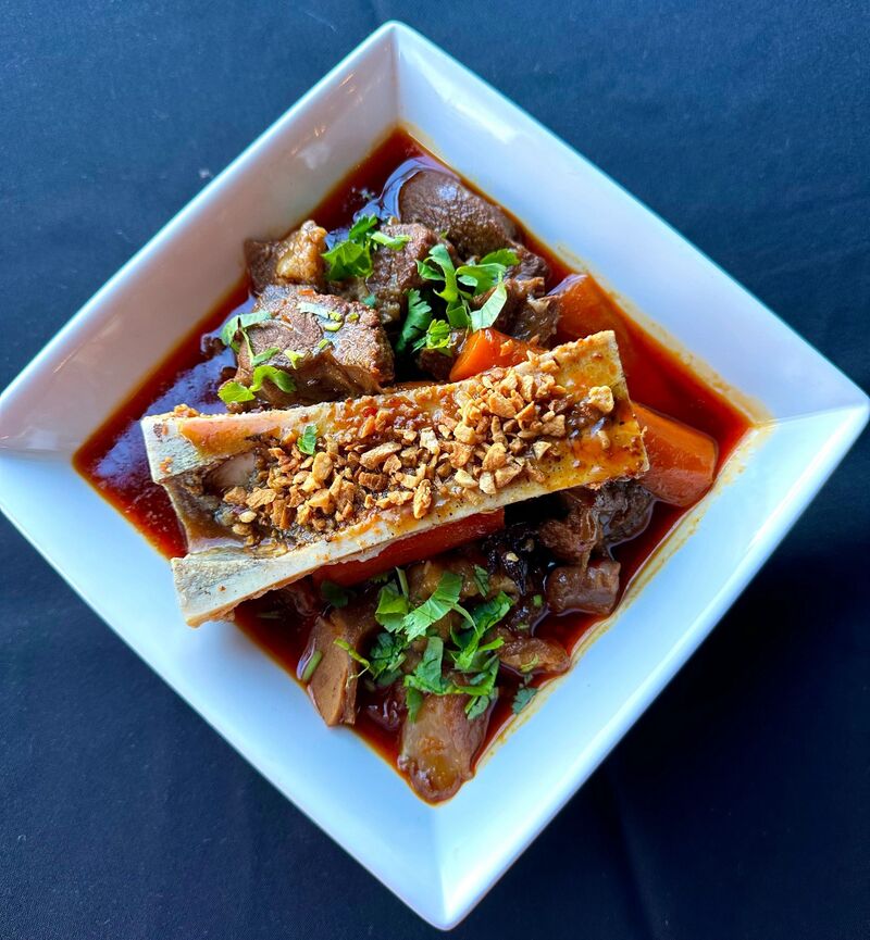 Overhead view of a square white bowl of braised beef and vegetables in red broth, topped with a halved bone and herbs.