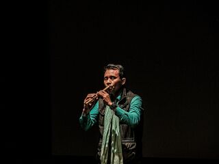 A man in a green shirt and scarf plays a flute alone on a dark stage.