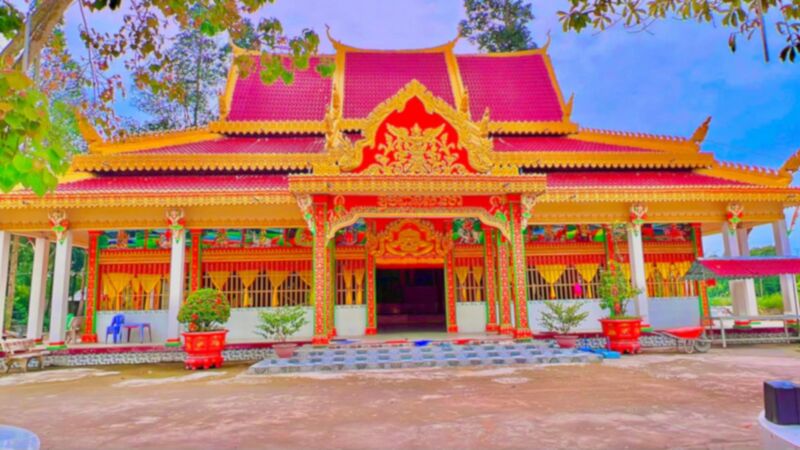 The front of a Khmer temple hall with a red tiered roof, gilded gable and red and gold columns.