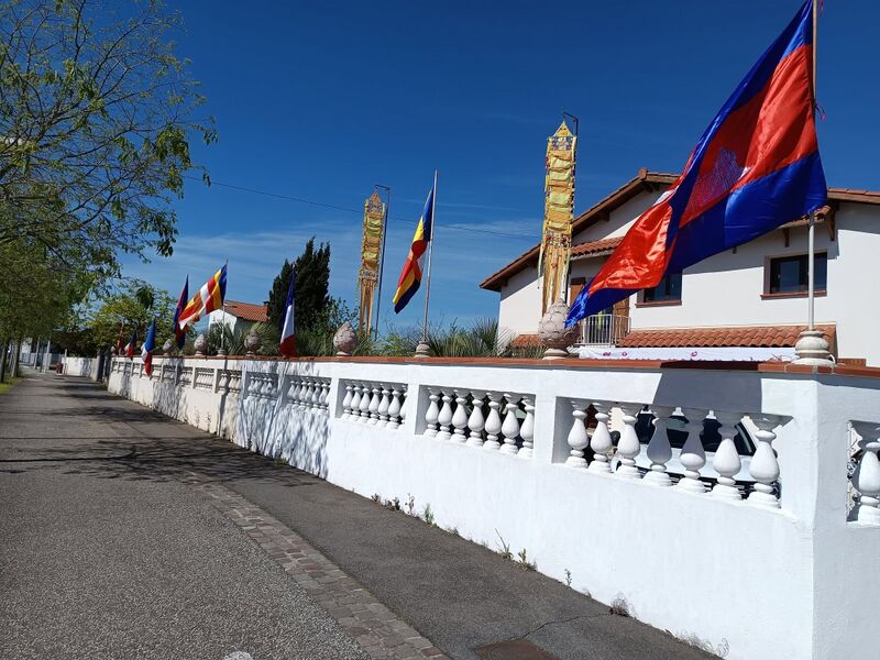 A white balustraded wall along a street with Buddhist and Cambodian flags and tall banners, a house behind.