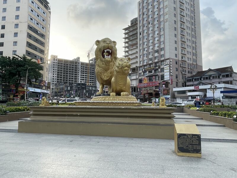 Two large gilded statues, a roaring maned lion and a lioness crouched in front of it, stand on a stepped plinth in the middle of a paved roundabout, with small gold lion figures at its corners and a black inscribed plaque in front. High-rise hotels, an unfinished concrete tower and a crane rise behind under a pale cloudy sky.
