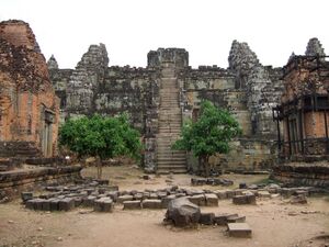 A steep stone stairway climbs the middle of a stepped pyramid of gray sandstone to a ruined shrine on top, with small towers standing on the tiers. Two leafy trees flank the foot of the stairs, loose blocks lie in rows on the dusty ground in front, and weathered brick towers stand at the left and right edges, one braced with wooden scaffolding.