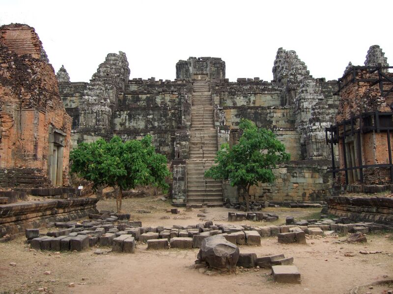 A steep stone stairway climbs the middle of a stepped pyramid of gray sandstone to a ruined shrine on top, with small towers standing on the tiers. Two leafy trees flank the foot of the stairs, loose blocks lie in rows on the dusty ground in front, and weathered brick towers stand at the left and right edges, one braced with wooden scaffolding.