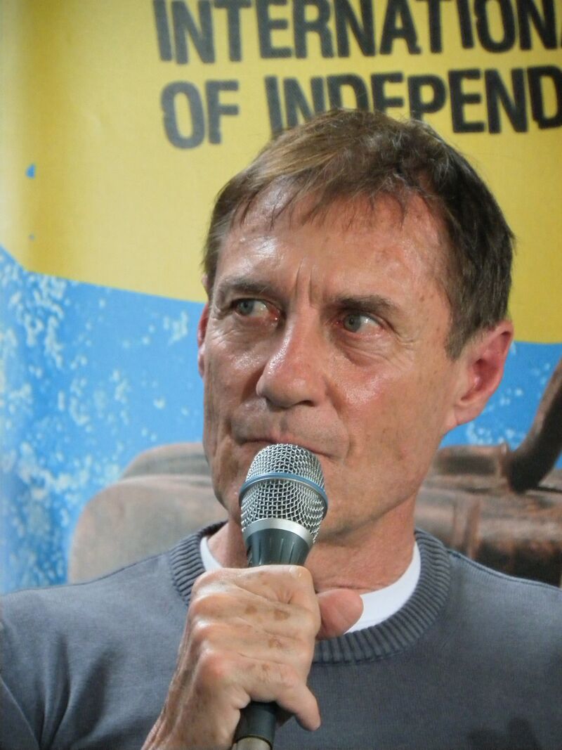 A grey-haired man speaking into a microphone in front of a yellow festival banner.