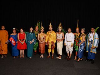 A row of people in Khmer dance costumes and formal clothes standing on a dark stage.