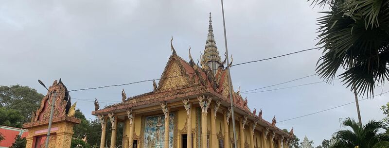 A Khmer temple hall with a tall tiered spire, ornate gables and a gilded gate, under a cloudy sky with palms.