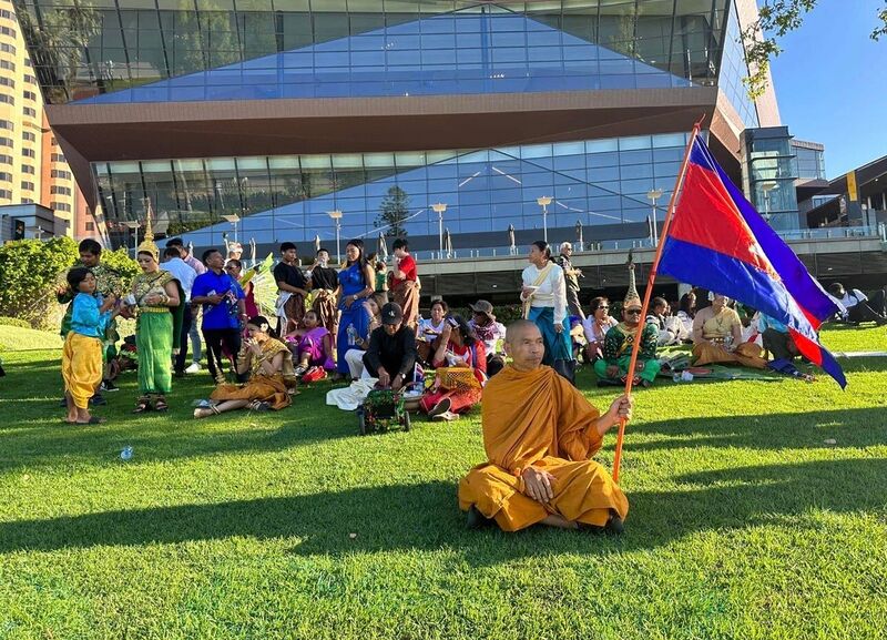 A monk sits on a lawn holding a Cambodian flag, with people in traditional dress gathered behind him outside a glass building.