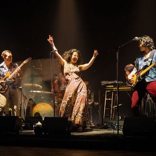 A singer in a long patterned dress raises her arms on stage between two guitarists and a keyboard player.