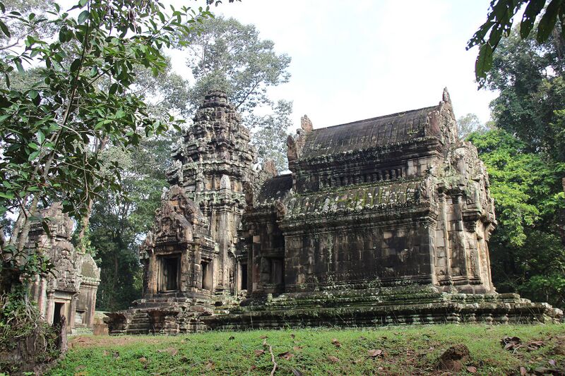 A carved sandstone sanctuary tower joined to a long hall, on a grassy base with tall trees around it.