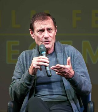A man in a grey jacket gestures while speaking into a microphone, seated before a green festival backdrop.