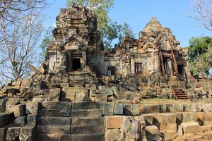 Two ruined stone towers joined by a low gallery with barred windows stand on a terrace of tumbled blocks under a clear blue sky. Red metal props hold up a doorway on the right, and large worn steps lead up in the foreground.