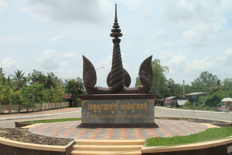 A tall dark monument with a spire and curved horns on a round paved plaza, with trees behind.