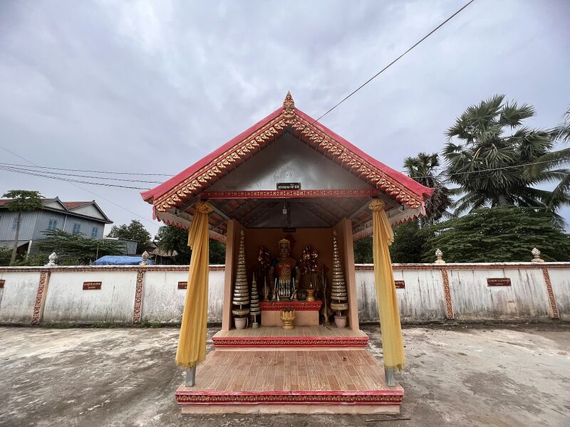 A small open shrine with a red tiled roof on yellow pillars, raised on a tiled platform, with a white wall and palm trees behind.
