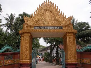 A tall gilded gateway with a pointed Khmer roof crest over a path leading into the temple grounds among palms.