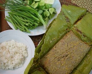 A banana-leaf parcel of grilled fish paste beside a plate of rice and a plate of green beans, cucumber and greens.