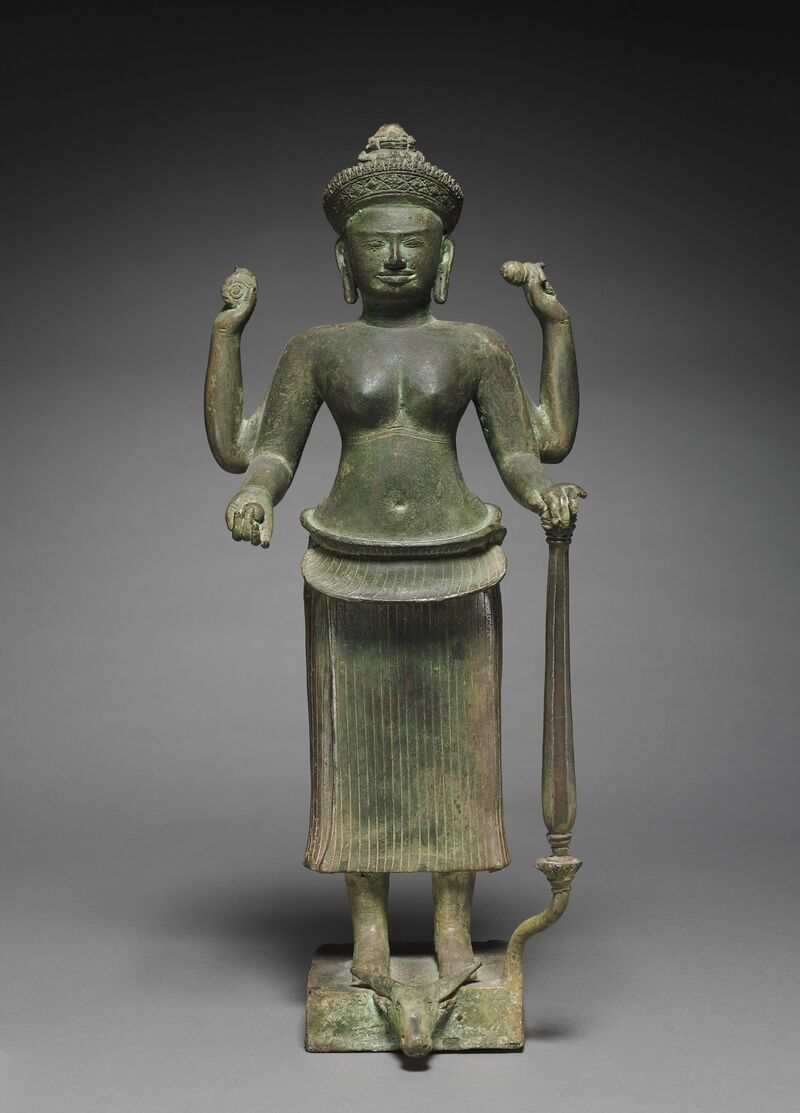 A dark green bronze goddess standing with four raised arms, wearing a tall crown and a long pleated skirt, one hand resting on a tall mace, with a buffalo head at her feet on a square base.