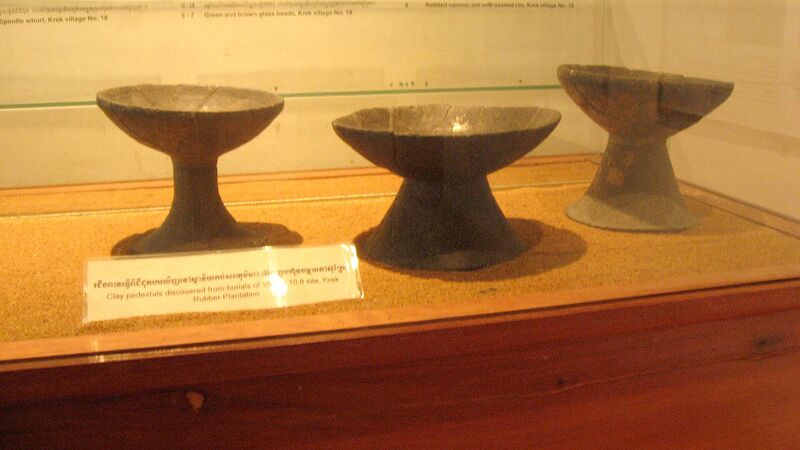 Three dark pottery bowls on tall feet in a glass display case, with a label in front.