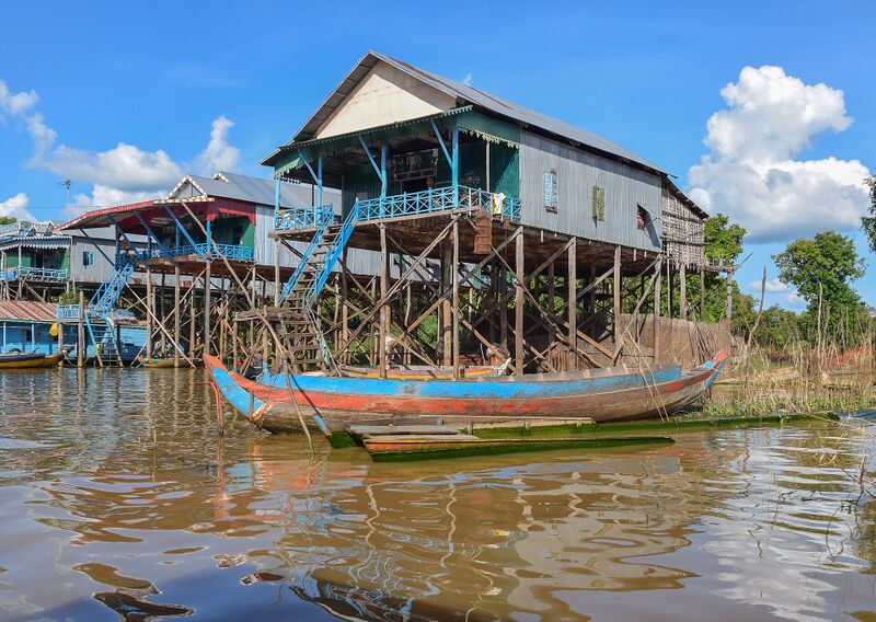 Tall wooden houses with metal roofs and blue railings stand on high stilts above brown water, with a long painted boat moored in front.