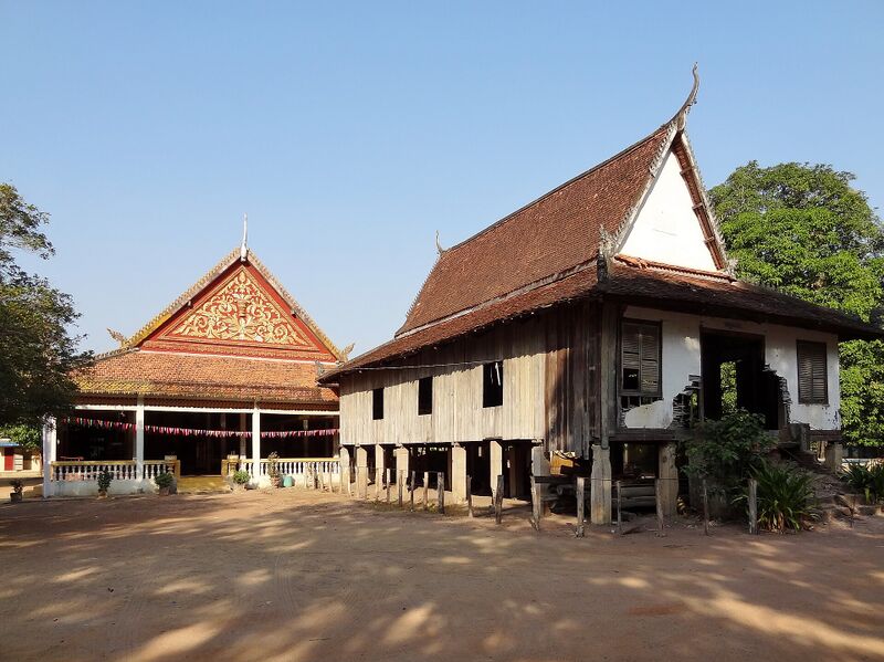 An open hall with a red and gold gable beside an old wooden hall on stilts with a steep tiled roof, on a sandy yard.