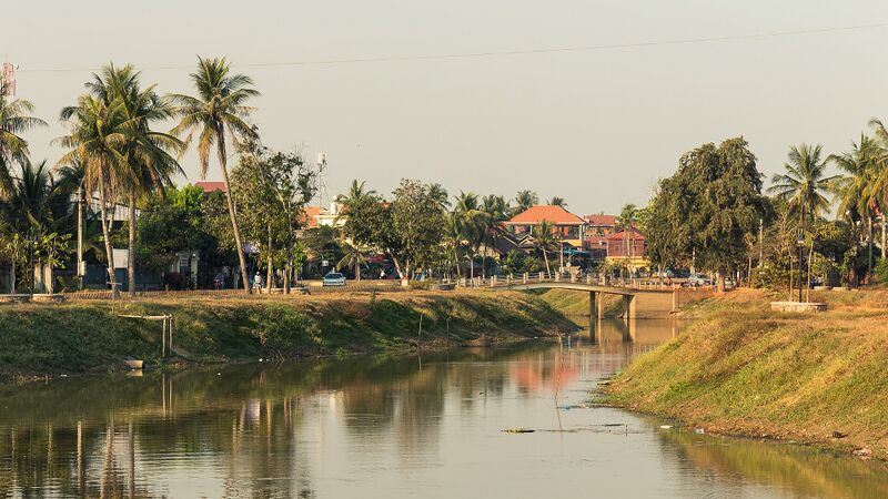 A calm river between grassy banks, crossed by a low bridge, with palm trees and tiled roofs beyond under a hazy sky.