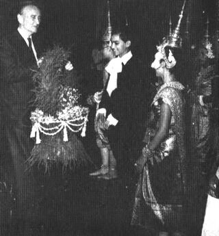 A man in a dark suit holds out a bouquet to a young woman in a tall dance crown and a young man in a suit.