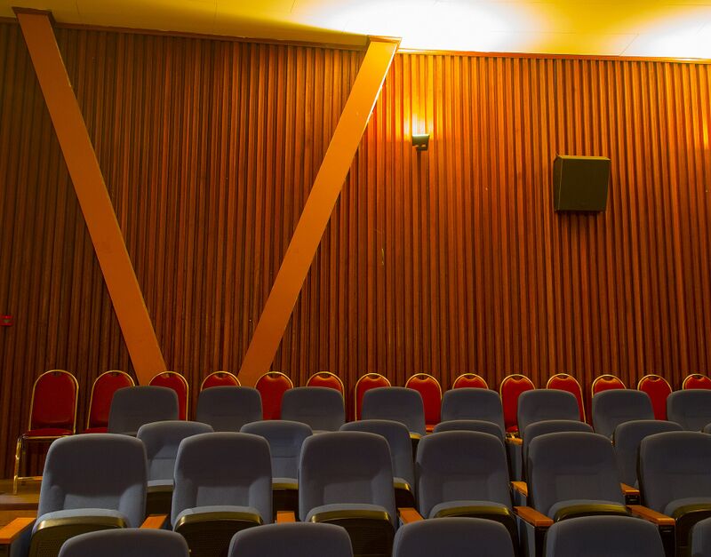 Rows of empty gray-blue theater seats face a wall of vertical warm-brown wooden slats crossed by two large orange beams in a V shape, with a row of red chairs behind the seats and a single wall lamp glowing.
