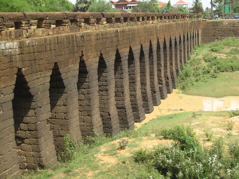 A long laterite bridge of tall narrow corbelled arches rising from a grassy bank, with green growth along its parapet.
