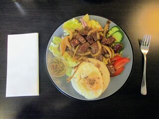 Seared beef strips with onion over salad and rice on a grey plate, a fried egg on the side, on a dark table.