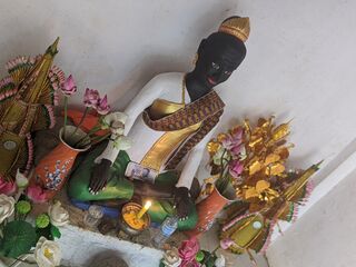 A reclining dark statue of a woman in a white blouse and gold sash, surrounded by flowers and offerings.