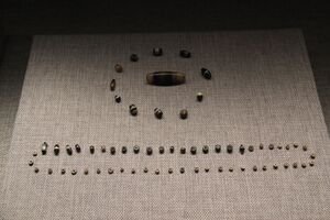 About seventy small beads laid out on gray linen in a museum case: a ring of banded dark and white stone beads around one long banded bead, and below them a looped line of tiny pale disc beads with a row of darker round beads along its top.