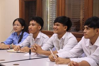 Four young students in white shirts sit at a table with notebooks, smiling during a lesson.