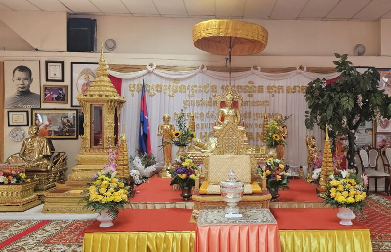 A shrine hall with gilded Buddha images, flowers, flags and a golden parasol on a red-carpeted platform.