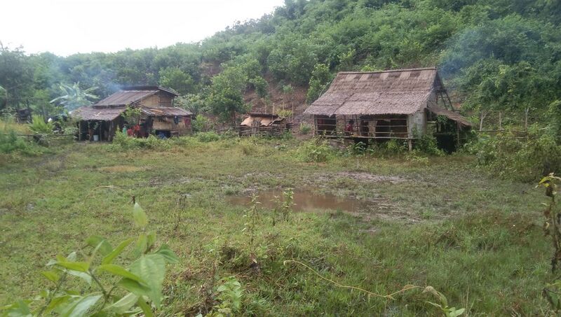 A cluster of small thatched wooden houses on a grassy hillside under a hazy sky, with forested hills behind