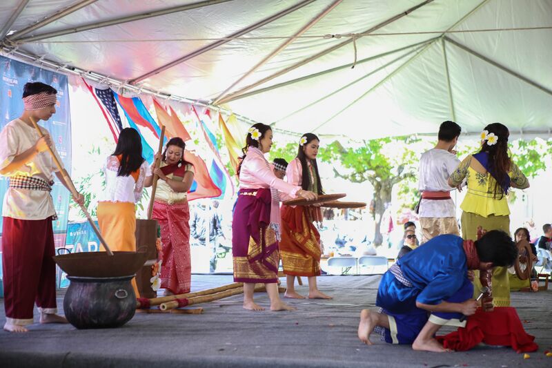 Performers in traditional Cambodian costume on a stage under a white tent, one kneeling, others standing.