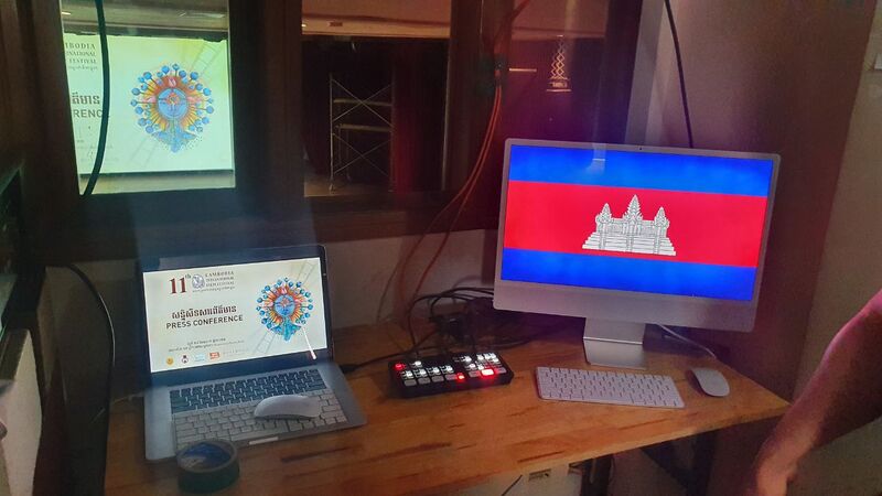 A laptop showing a press conference slide for the 11th Cambodia International Film Festival beside a monitor showing the Cambodian flag, in a dim projection booth.