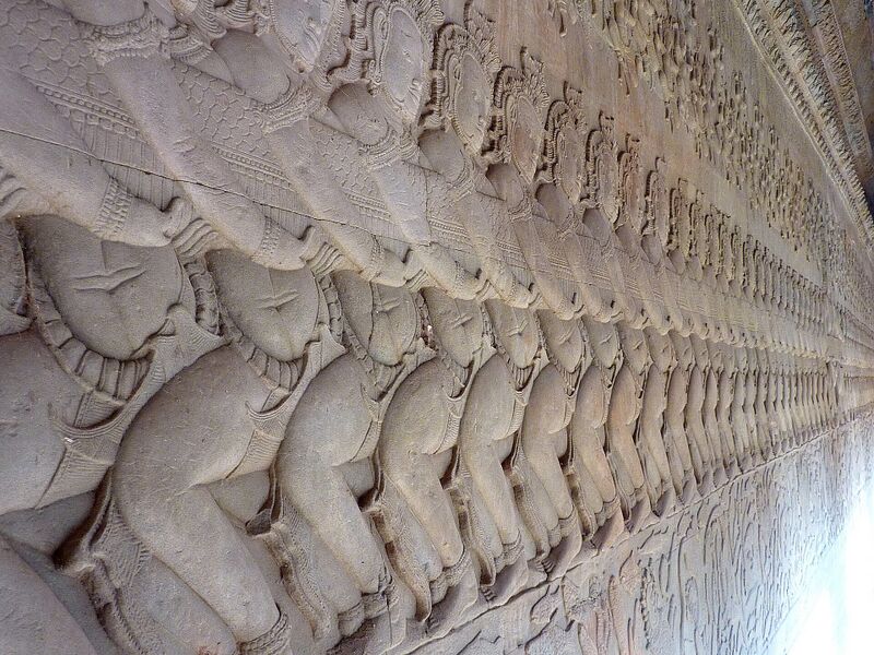 A long carved stone wall in which a row of figures in profile pulls together on the body of a serpent, receding into the distance.